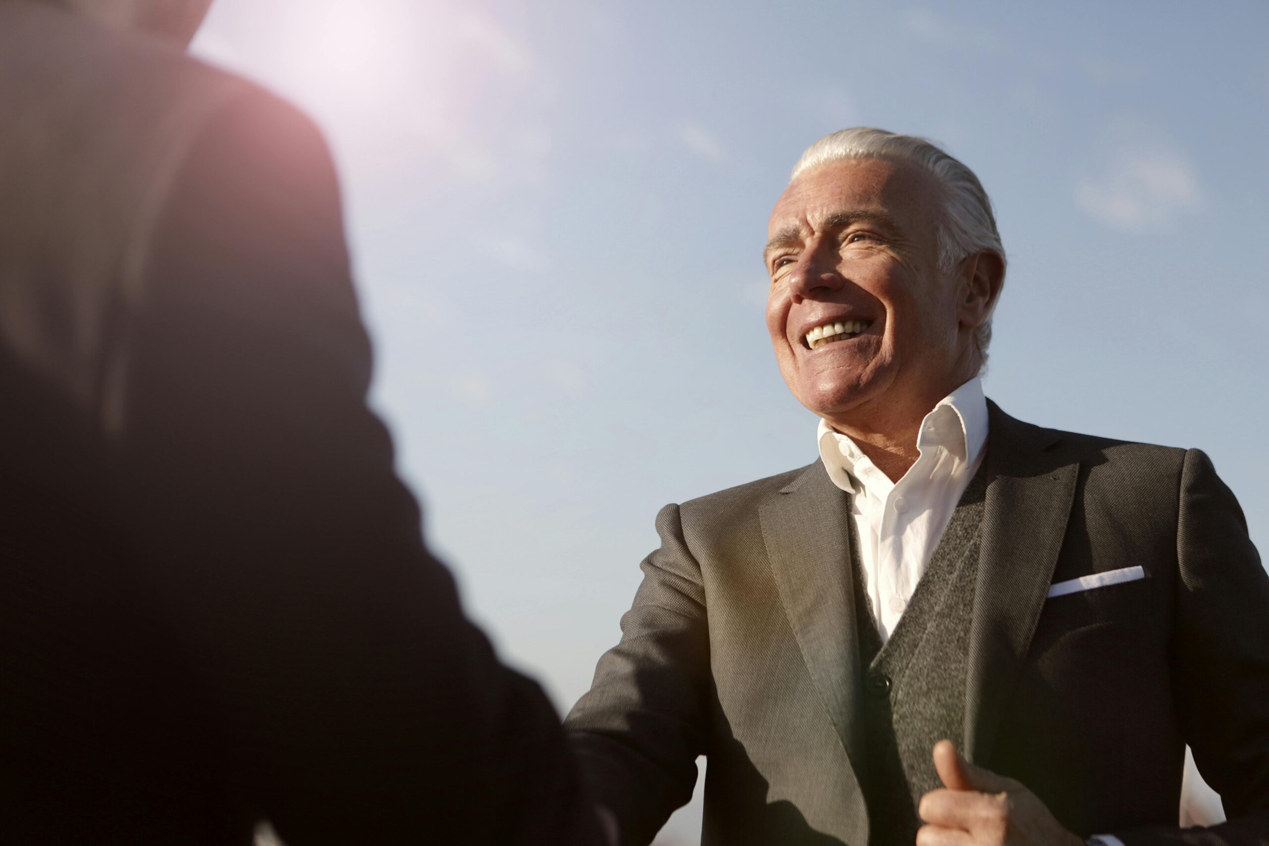 Elderly man in formal wear smiling while shaking hands outside during the day.